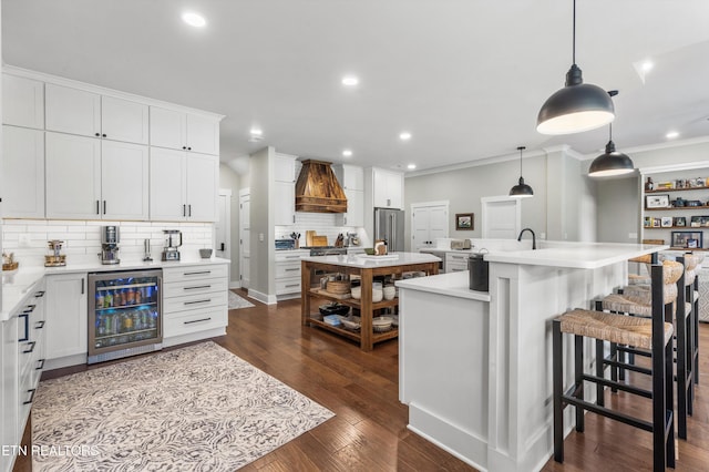 kitchen with custom range hood, pendant lighting, white cabinets, wine cooler, and an island with sink