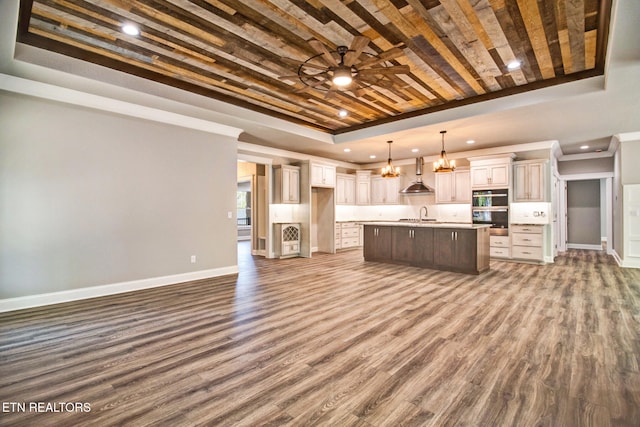 kitchen featuring an island with sink, a tray ceiling, dark hardwood / wood-style floors, pendant lighting, and wall chimney exhaust hood