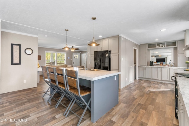 kitchen featuring wood-type flooring, a kitchen bar, black fridge with ice dispenser, decorative light fixtures, and a kitchen island with sink