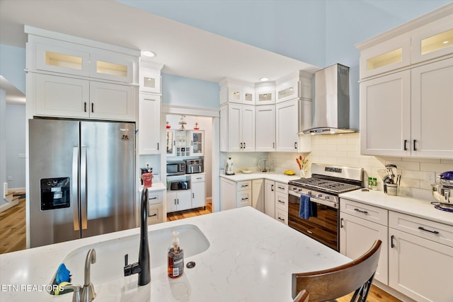 kitchen featuring wall chimney exhaust hood, white cabinets, light stone countertops, and stainless steel appliances