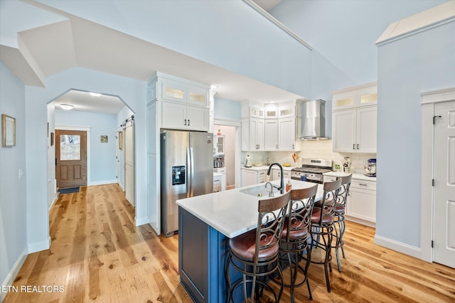 kitchen with light wood-type flooring, an island with sink, white cabinetry, stainless steel appliances, and wall chimney exhaust hood