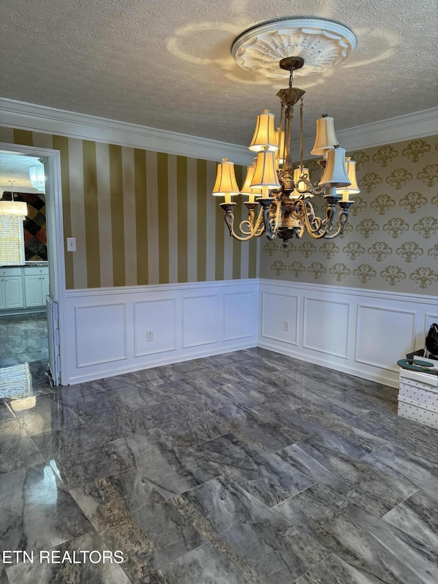 unfurnished dining area with wallpapered walls, a wainscoted wall, ornamental molding, a textured ceiling, and a chandelier