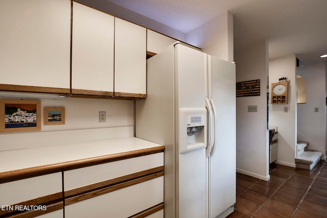 kitchen featuring white refrigerator with ice dispenser, light countertops, white cabinetry, dark tile patterned floors, and baseboards