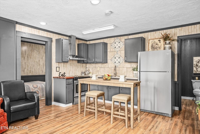 kitchen with crown molding, stainless steel appliances, wall chimney range hood, and light wood-type flooring