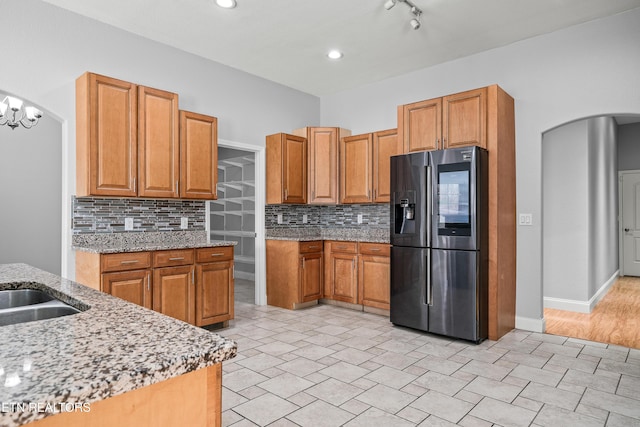 kitchen with brown cabinetry, decorative backsplash, stainless steel fridge, arched walkways, and light stone countertops