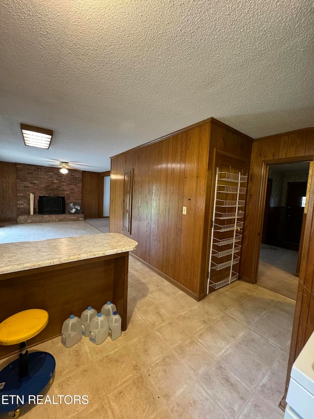 kitchen with a brick fireplace, ceiling fan, wooden walls, and a textured ceiling