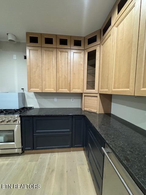 kitchen featuring light brown cabinets, appliances with stainless steel finishes, dark stone counters, light wood finished floors, and glass insert cabinets