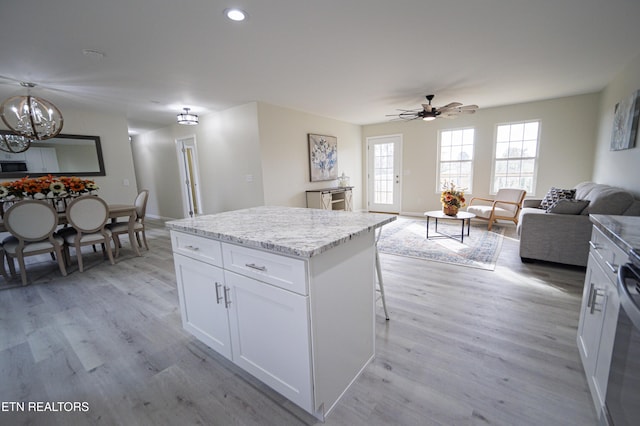 kitchen with light stone countertops, ceiling fan with notable chandelier, a kitchen island, white cabinets, and light wood-type flooring