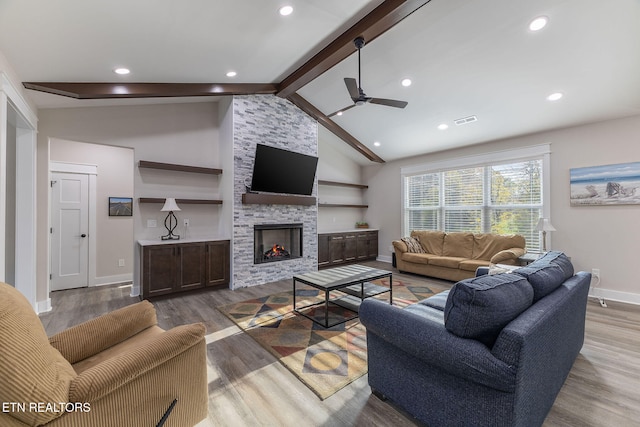 living room with a stone fireplace, ceiling fan, wood-type flooring, and vaulted ceiling with beams