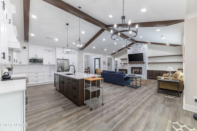 kitchen featuring a stone fireplace, stainless steel appliances, a center island with sink, sink, and white cabinetry