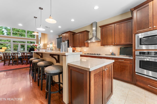 kitchen with wall chimney exhaust hood, a breakfast bar area, decorative light fixtures, a kitchen island, and light stone countertops