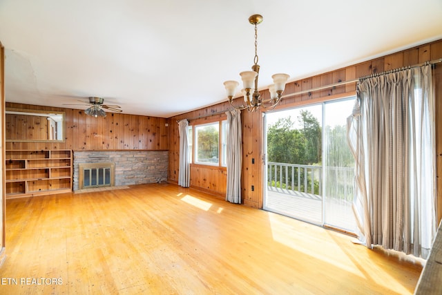 unfurnished living room with wood walls, a stone fireplace, ceiling fan with notable chandelier, and hardwood / wood-style floors