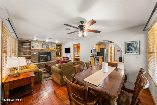 dining area featuring a stone fireplace, hardwood / wood-style floors, built in features, and ceiling fan