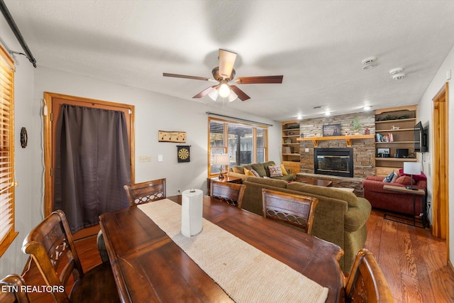 dining space featuring built in shelves, a stone fireplace, a textured ceiling, dark wood-type flooring, and ceiling fan