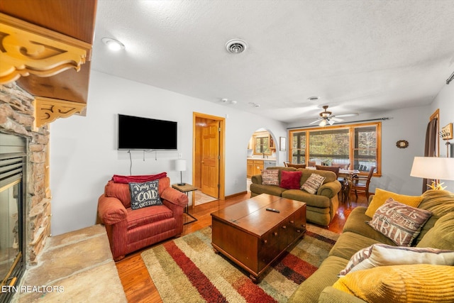 living room featuring hardwood / wood-style floors, a stone fireplace, a textured ceiling, and ceiling fan