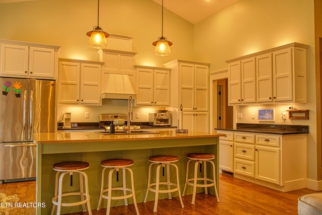 kitchen featuring pendant lighting, a kitchen island with sink, high vaulted ceiling, sink, and stainless steel refrigerator