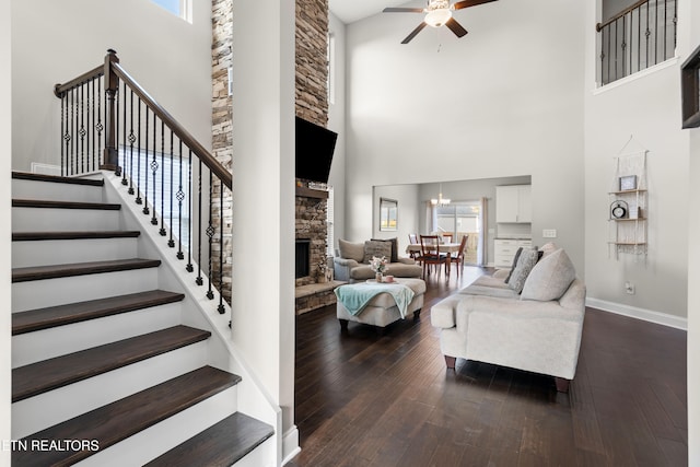 living room featuring a stone fireplace, ceiling fan with notable chandelier, dark wood-type flooring, and a high ceiling