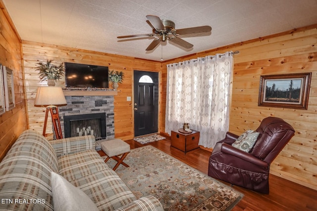 living room with a stone fireplace, wooden walls, wood-type flooring, and ceiling fan