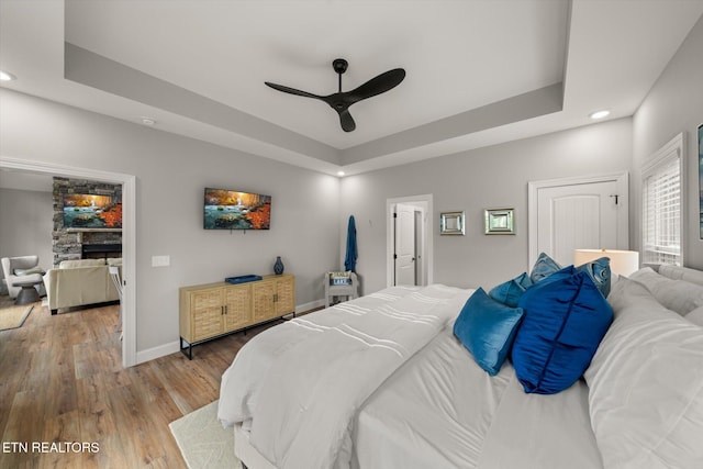 bedroom with hardwood / wood-style floors, a stone fireplace, a raised ceiling, and ceiling fan