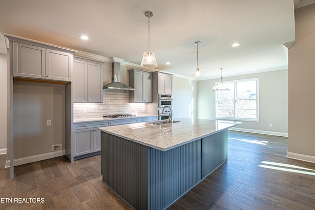 kitchen featuring pendant lighting, stainless steel gas stovetop, a center island with sink, wall chimney range hood, and light stone countertops