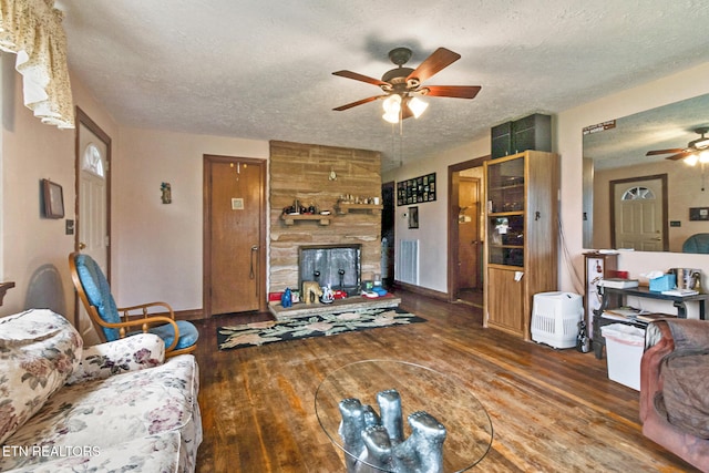 living room featuring a textured ceiling, ceiling fan, a stone fireplace, and dark wood-type flooring