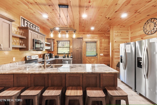 kitchen featuring kitchen peninsula, light stone counters, stainless steel appliances, hanging light fixtures, and wood walls