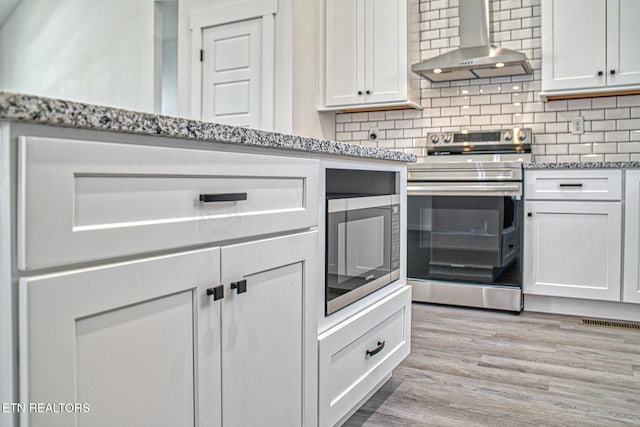 kitchen featuring white cabinetry, stainless steel appliances, wall chimney range hood, light stone counters, and light hardwood / wood-style flooring