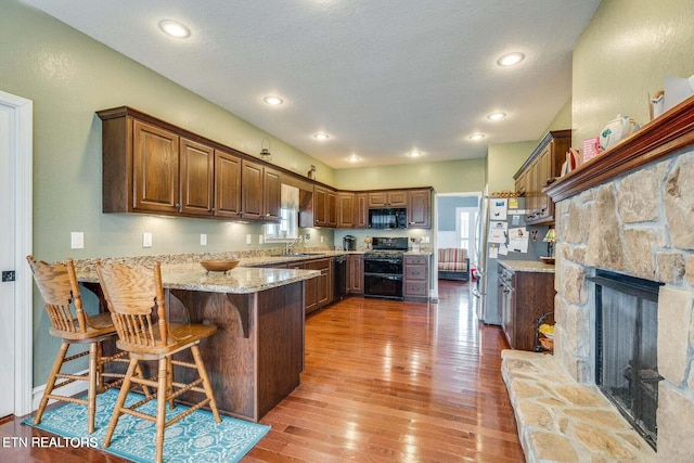 kitchen with black appliances, a kitchen breakfast bar, a stone fireplace, hardwood / wood-style flooring, and kitchen peninsula