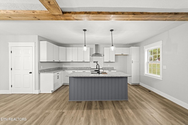 kitchen featuring beam ceiling, a center island with sink, white cabinets, and wall chimney range hood