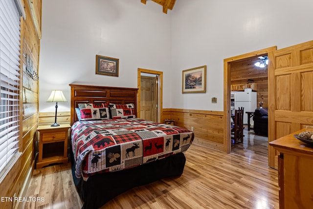 bedroom featuring wood walls, light hardwood / wood-style flooring, a towering ceiling, and white fridge