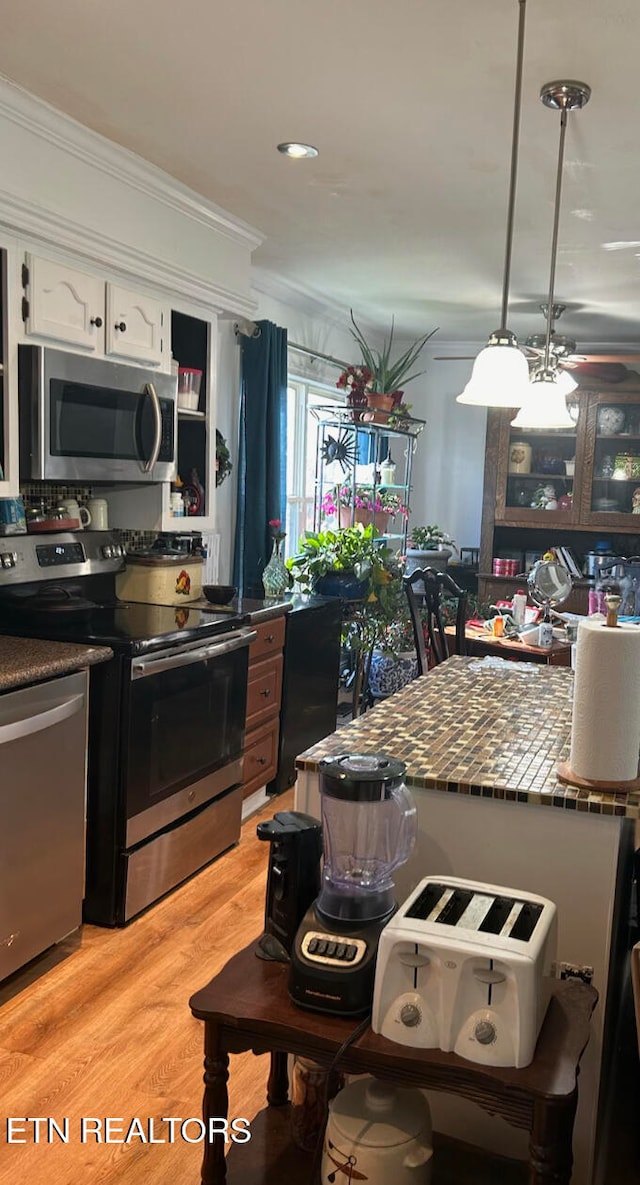 kitchen with light wood-type flooring, ornamental molding, stainless steel appliances, decorative light fixtures, and white cabinets