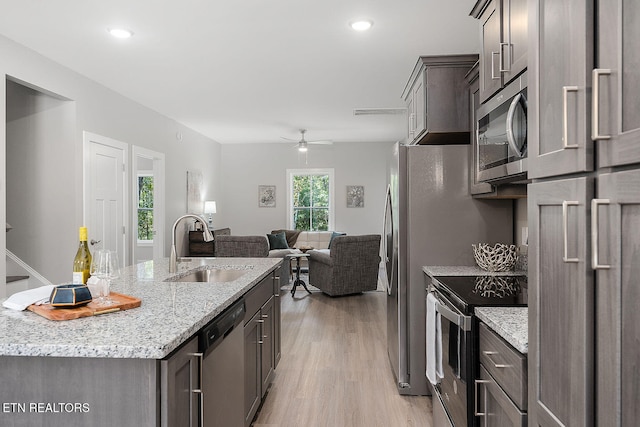 kitchen with sink, stainless steel appliances, light stone counters, an island with sink, and light hardwood / wood-style floors