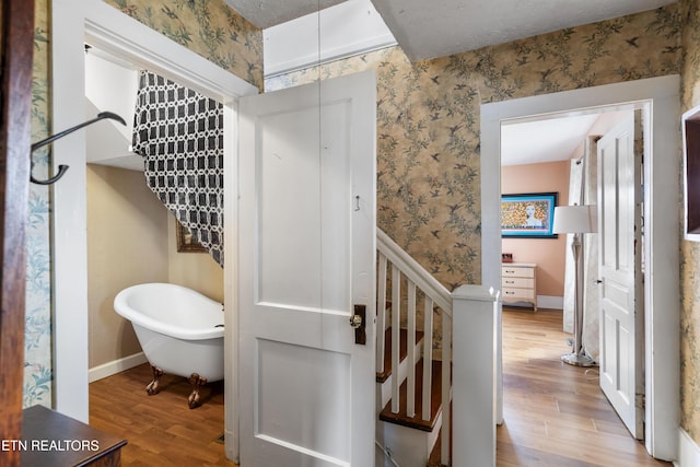 bathroom featuring a textured ceiling, wood finished floors, baseboards, a freestanding bath, and wallpapered walls