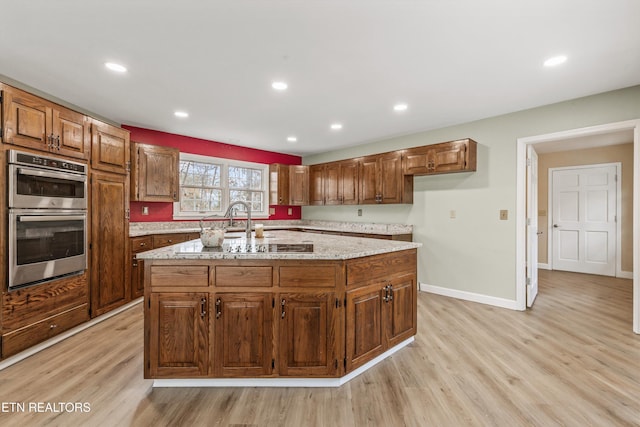 kitchen with light stone counters, light wood-type flooring, stainless steel double oven, and a kitchen island with sink