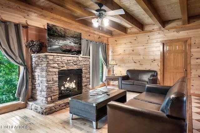 living room with beam ceiling, ceiling fan, a stone fireplace, light hardwood / wood-style floors, and wooden walls