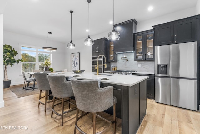 kitchen with stainless steel refrigerator with ice dispenser, custom range hood, sink, a center island with sink, and hanging light fixtures