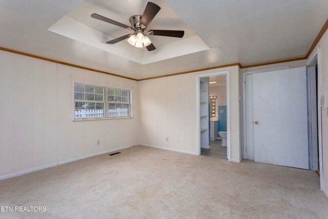 unfurnished bedroom with ensuite bath, ornamental molding, light colored carpet, a tray ceiling, and ceiling fan