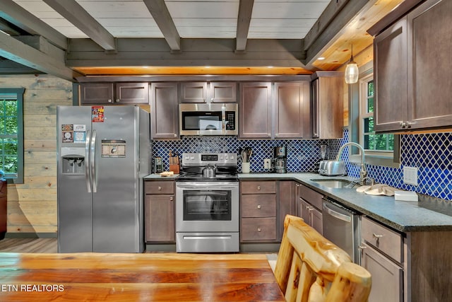 kitchen featuring stainless steel appliances, wooden walls, sink, beam ceiling, and decorative light fixtures