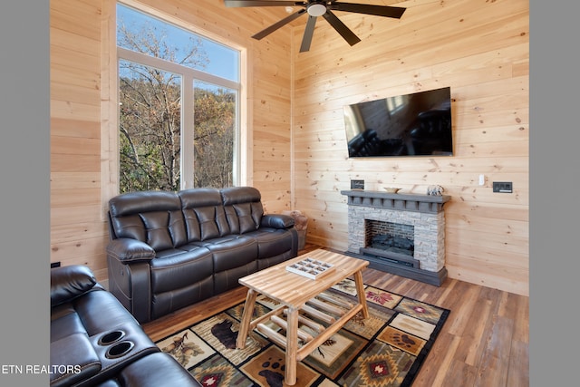 living room featuring hardwood / wood-style flooring, a stone fireplace, ceiling fan, and wooden walls