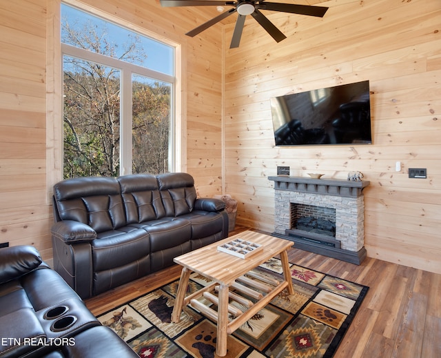 living room featuring hardwood / wood-style flooring, a stone fireplace, ceiling fan, and wood walls