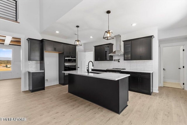 kitchen with tasteful backsplash, wall chimney exhaust hood, a kitchen island with sink, sink, and light hardwood / wood-style floors