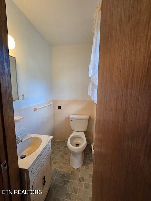 bathroom with toilet, a wainscoted wall, stone finish flooring, vanity, and tile walls