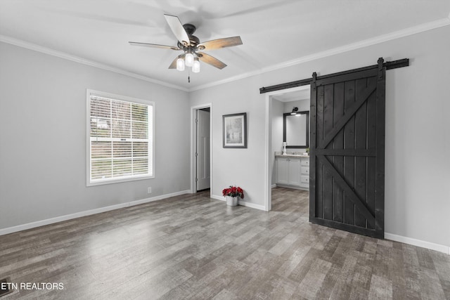 unfurnished bedroom featuring hardwood / wood-style floors, ensuite bathroom, crown molding, ceiling fan, and a barn door