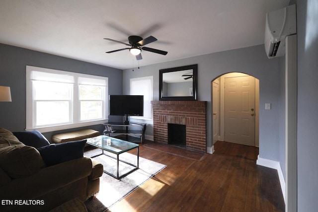 living room with a brick fireplace, ceiling fan, dark wood-type flooring, and a wall unit AC