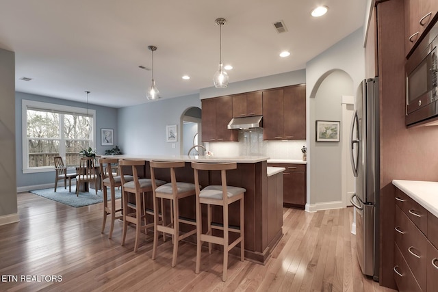 kitchen with pendant lighting, light hardwood / wood-style flooring, a kitchen island with sink, backsplash, and stainless steel appliances