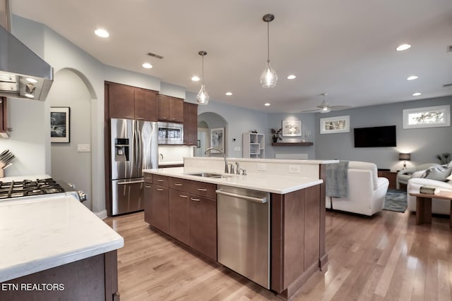 kitchen featuring sink, hanging light fixtures, an island with sink, stainless steel appliances, and wall chimney range hood