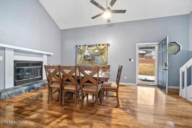 dining room with hardwood / wood-style floors, ceiling fan, a stone fireplace, and high vaulted ceiling