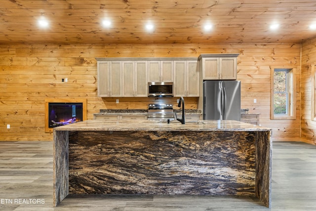 kitchen with light stone countertops, appliances with stainless steel finishes, wooden walls, wood-type flooring, and an island with sink