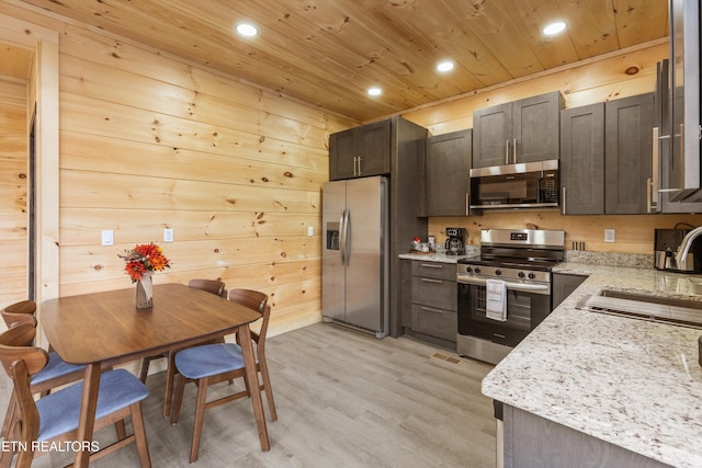 kitchen featuring appliances with stainless steel finishes, dark brown cabinets, wooden walls, and light stone counters