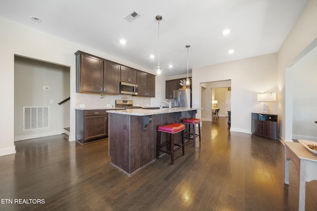 kitchen featuring a kitchen bar, stainless steel appliances, a kitchen island with sink, hanging light fixtures, and light stone countertops